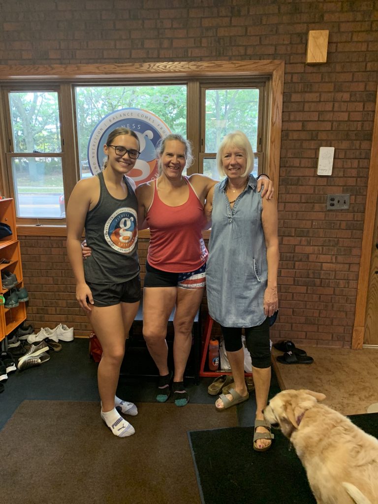 three women (one older, one middle age, one younger) stand in a gym entry way wearing work out clothing. a dog is in the foreground.