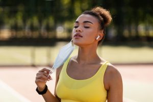 Portrait of African American young woman taking off protective medical mask and breathing fresh air, wearing wireless headphones exercising outside in the city during covid epidemic outbreak